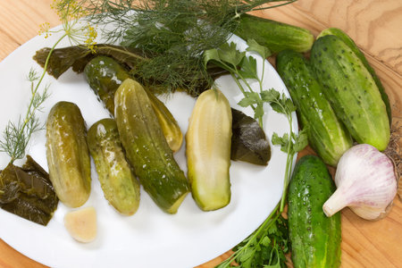 Whole and halved homemade pickled cucumbers among the some herbs and spices for their pickling on a dish and fresh cucumbers, fragment close-upの写真素材