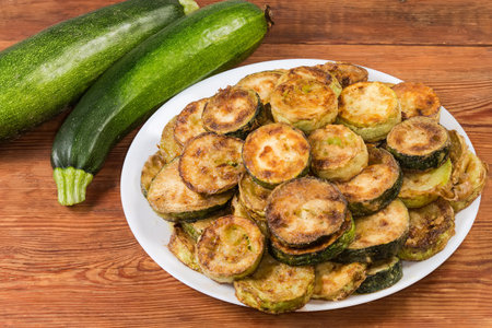 Heap of fried green zucchini and yellow vegetable marrows sliced into circles on the white dish against the whole raw green zucchini on a rustic tableの写真素材