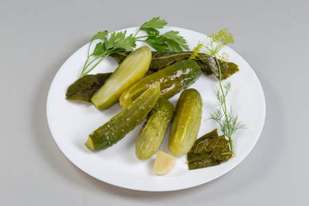 Several whole and one halved homemade pickled cucumbers among the some herbs and spices for their pickling on a white dish on a gray backgroundの写真素材