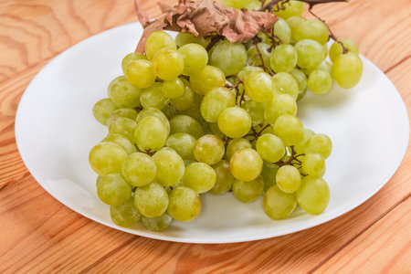 Clusters of the ripe white sultana grape with dry vine leaf on a white dish on a wooden rustic table, side view close-up in selective focusの写真素材