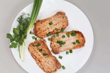 Grilled chops made from boneless slices of pork loin with fresh green onion, dill and parsley on the white dish, top view on a gray backgroundの写真素材