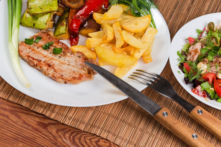 Serving of grilled boneless pork chop with the baked vegetables and French fries on the white dish and tuna salad on a rustic table close-upの写真素材
