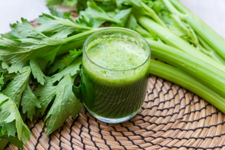 Glass of freshly squeezed celery juice against the fresh stems of celery with leaves on the table with table mat, close-up in selective focusの写真素材
