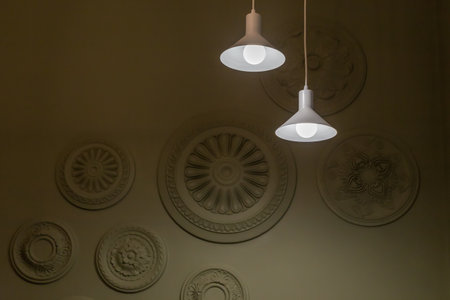 Two glowing pendant lights with LED lamps and white simple metallic conical bodies suspended from the ceiling by the long power cords on a dark background of wall with decorationsの写真素材