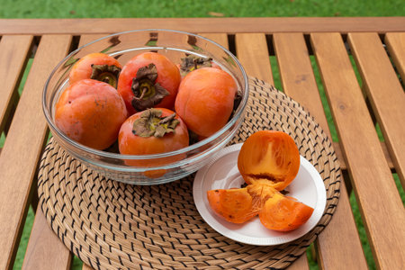 Ripe whole persimmon fruits in the transparent glass kitchen bowl and one fruit cut into three parts on a saucer on wooden table with place mat outdoorsの写真素材