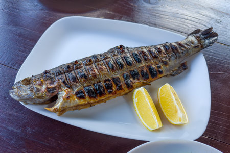 Whole grilled river trout with fresh lemon slices on the white dish on a wooden table
