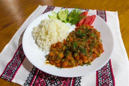 Portion of boiled rice, served with stewed vegetables mix and slices of fresh vegetables on the white dish with napkin on a wooden tableの写真素材