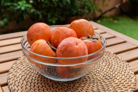 Ripe persimmon fruits in the transparent glass kitchen bowl on wooden table with place mat outdoorsの写真素材