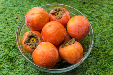 Ripe persimmon fruits in the transparent glass kitchen bowl standing on a grass, top viewの写真素材