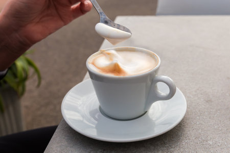Cappuccino with a foam layer in white cup with handle standing on saucer and milk foam in spoon in a...
