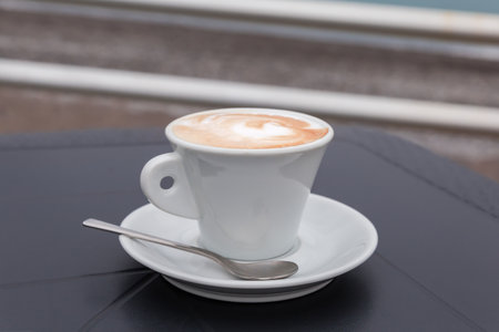 Cappuccino with drawing on a foam layer atop in white cup standing on a saucer with tea spoon on a black tableの写真素材