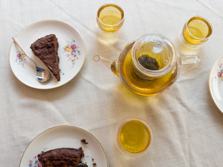 Freshly brewed green tea with herbal mix in three transparent glass cups and in the glass teapot with cake pieces on saucers on a table covered with tablecloth, top viewの写真素材