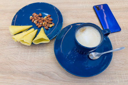 Coffee with milk prepared with capsule coffee maker in blue ceramic cup, served with cheese and nuts, unused single-serve coffee capsules on the wooden tableの写真素材