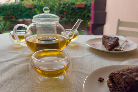 Brewed green tea in glass cup against the other the same cups and glass teapot and cake pieces on saucers on a table covered with tablecloth, front view close-up in selective focusの写真素材