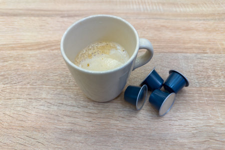 Coffee with milk in white ceramic cup prepared with capsule coffee maker and several unused single-serve coffee capsules on the wooden table, close-up in selective focusの写真素材