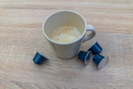 Coffee with milk in white ceramic cup prepared with capsule coffee maker and several unused single-serve coffee capsules on the wooden table, close-up in selective focusの写真素材