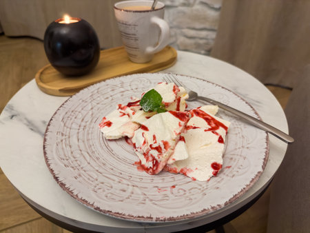 Dessert of meringue and mascarpone, poured over with raspberry jam on dish on a small round table against the coffee cup and burning candleの写真素材
