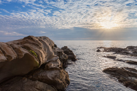 Part of the sea shore with stone coastline on a background of the partly cloudy sky and sun beams...