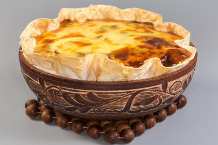 Hot baked sweet cottage cheese casserole in flat bread in a clay bowl on a wooden hot stand woven from wooden beads on a gray background, front view in selective focusの写真素材
