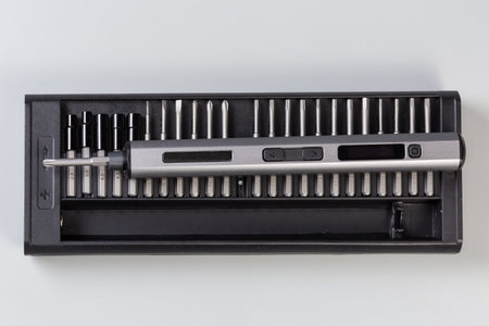 Electric rechargeable cordless mini screwdriver in metal tubular housing lies atop on the set of different interchangeable bits in plastic case, top view on a gray backgroundの写真素材