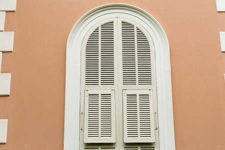 Upper part of the arched window in yellow wall with closed wooden shutters painted white, view outdoors in overcast dayの写真素材
