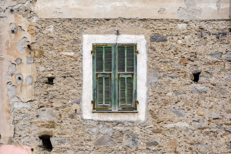 Fragment of an ancient rough stone wall with window covered with green wooden exterior shutters with fixed louvers and rusty shutter hardware in overcast dayの写真素材