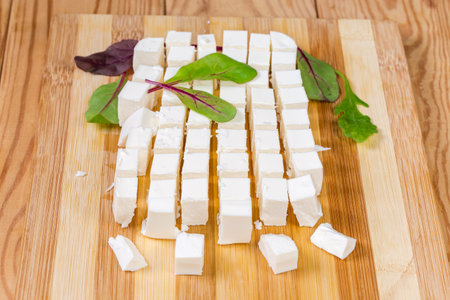 Diced piece of soft cheese with leaves of fresh herbs on the wooden bamboo cutting board on a rustic tableの写真素材