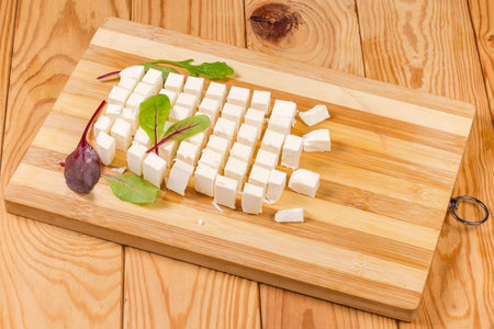 Diced piece of soft cheese with leaves of fresh herbs on the wooden bamboo cutting board on a rustic tableの写真素材