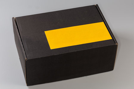 Closed rectangular packing box made with black cardboard with blank yellow sticker atop on a gray backgroundの写真素材