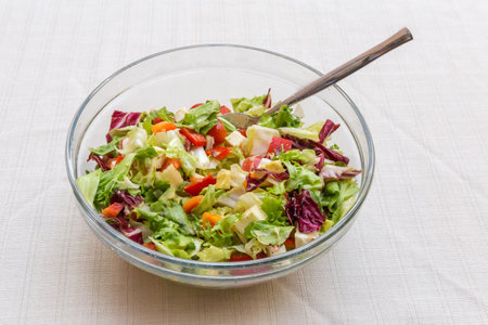 Salad of diced mozzarella, fresh vegetables and different type of leaf lettuce in a glass bowl on a table covered with white tableclothの写真素材