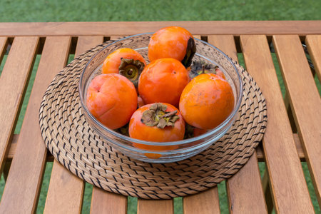 Ripe organic persimmon fruits in the transparent glass kitchen bowl on a wooden table with place mat, outdoors in patio in overcastの写真素材