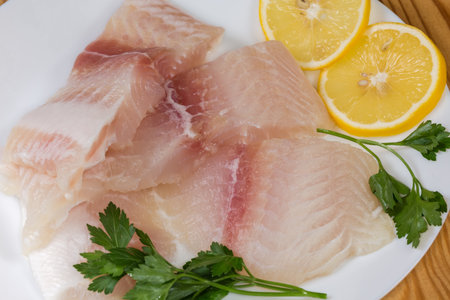 Raw pieces of the defrosted fillet of the Pangasius, also known as basa fish on a dish with lemon slices and fresh parsley twigs, top view close-upの写真素材
