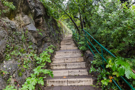 Fragment of wet paved stone staircase with metal railing painted green next to a precipitous rock on...