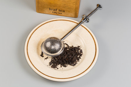 Closed spherical stainless steel tea-strainer on a saucer with dried black tea leaves against the wooden tea box on a gray background, top viewの写真素材