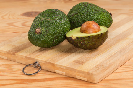 Two whole and one half of avocado with seed and dark green skin on a bamboo wooden cutting board on a rustic table, front view close-upの写真素材