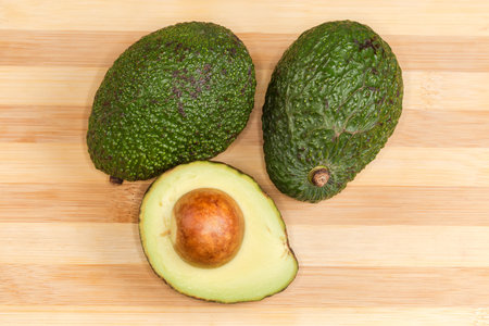 Two whole and one half of avocado with seed and dark green skin on a bamboo wooden cutting board, top view close-upの写真素材