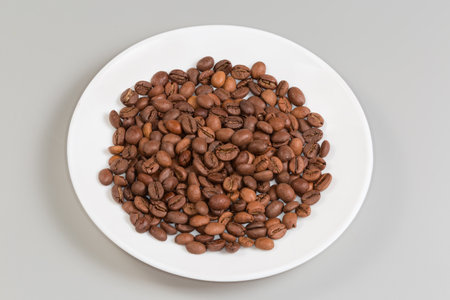 Pile of roasted coffee beans light brown of medium degrees of roast on a white dish on gray backgroundの写真素材