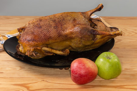 Baked whole carcass of the domestic duck stuffed with apples on a big black plate on the rustic table, front view with fresh apples on a foregroundの写真素材