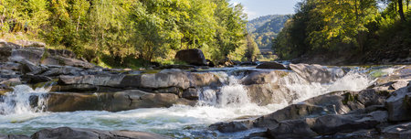 Multi-level water cascade waterfall on a mountain river among the forest on slopes in autumn sunny day, panoramic view from a lower shooting pointの写真素材