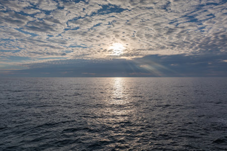 Section of the sky with altocumulus clouds over the water surface of sea with sunny path and distant storm clouds on a horizon at autumn morning backlitの写真素材