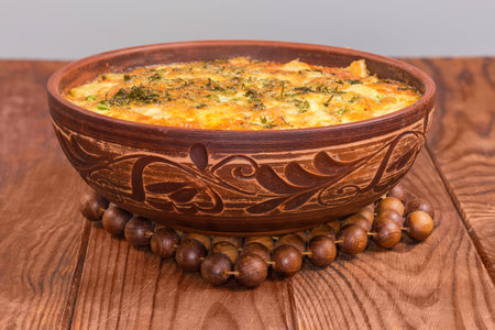 Freshly prepared potato casserole with browned crust of melted cheese in clay bowl on wooden hot stand on a rustic table, front view close-up in selective focusの写真素材