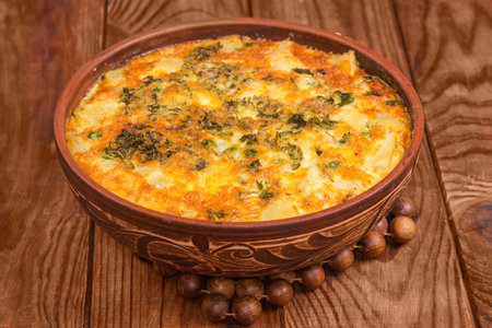 Freshly prepared potato casserole with appetizing browned crust of melted cheese on the top in clay bowl on wooden hot stand on a rustic table, front view close-upの写真素材