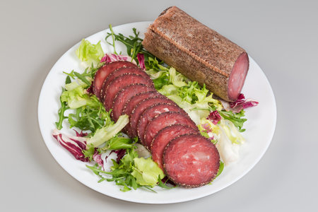 Slices of the dry-cured sausage covered with spices on a fresh leaf vegetables and piece of the same sausage in the textile casing on dish on a gray backgroundの写真素材
