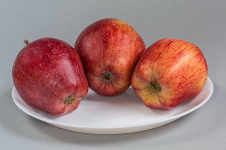 Three whole fresh red apples oblong shape on the white plate on a gray background, front view close-upの写真素材