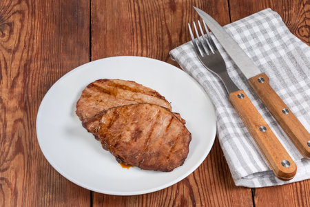 Serving of two pork loin steaks cooked on grill on a white plate and cutlery on an old rustic tableの写真素材