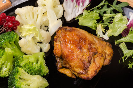 Crispy baked chicken thigh with boiled broccoli and cauliflower pieces and fresh leaf vegetables on the black dish, fragment top viewの写真素材