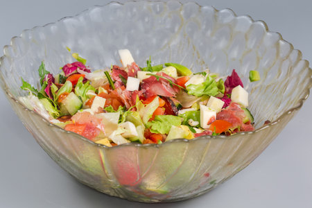 Salad with soft cheese, chopped dry-cured sausage, avocado and different green and red lettuce, other fresh vegetables in glass salad bowl, front view close-up in selective focusの写真素材