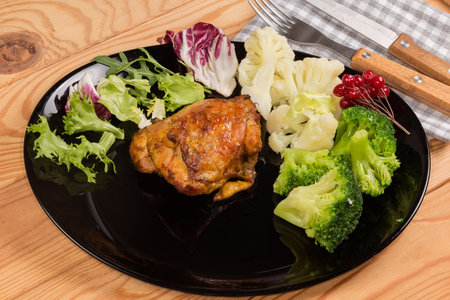 Serving of crispy baked chicken thigh with boiled broccoli and cauliflower pieces and fresh leaf vegetables on the black dish on a rustic tableの写真素材
