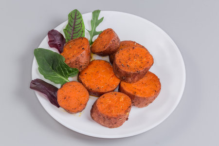Portion of the baked circle slices of the sweet potato tubers in their skin served with greens leaves on the dish on a gray backgroundの写真素材
