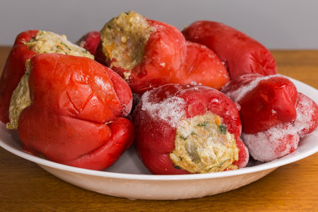 Frozen raw red bell peppers stuffed with rice and chopped meat for following final cooking lie on a dish on a wooden table, front view close-up in selective focusの写真素材